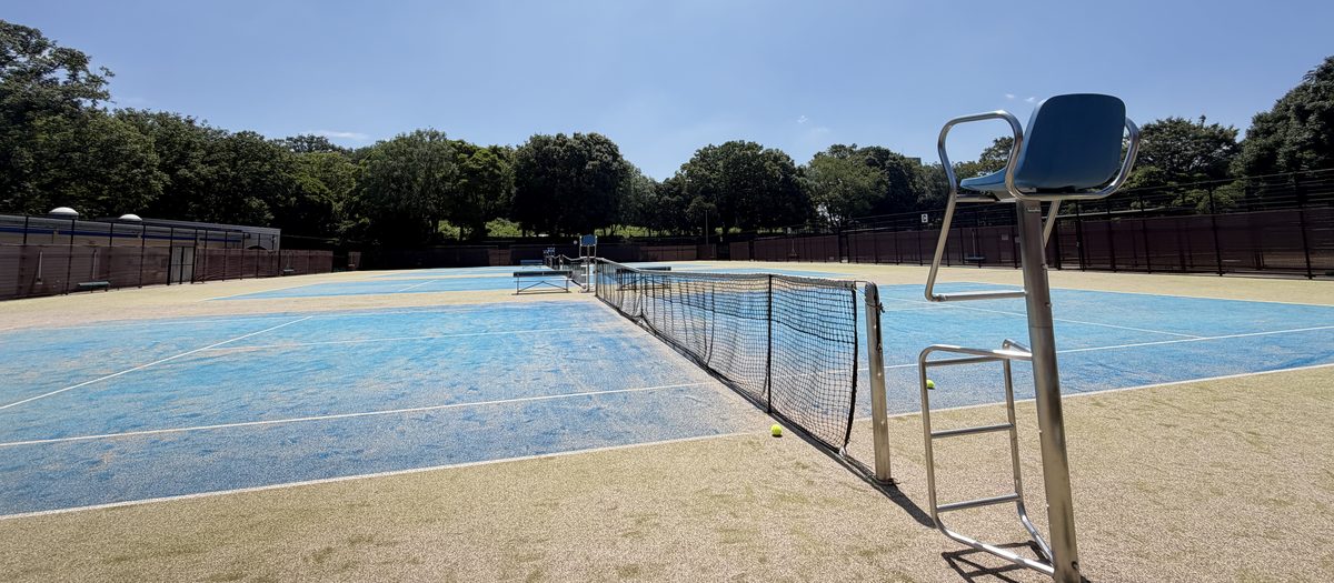 An outdoor omni court in western Tokyo — sand-filled artificial grass, a blue inner court, and a line of trees behind the fence.
